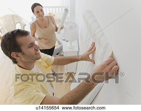 Picture - Pregnant Hispanic couple hanging wall paper in nursery. Fotosearch