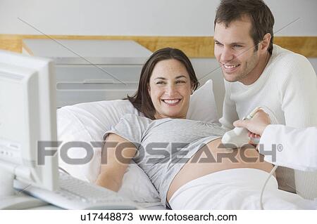 Stock Image - Pregnant Hispanic couple looking at ultrasound monitor. Fotosearch