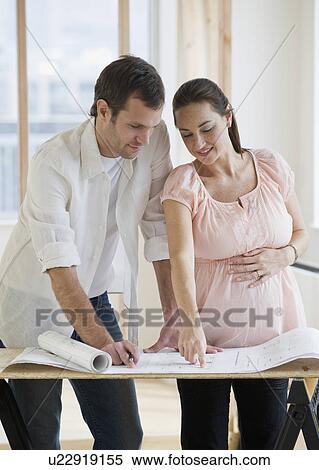 Stock Photography - Pregnant Hispanic couple looking at blueprints. Fotosearch