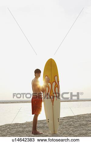 Stock Image - Teenage boy standing with surfboard on beach. Fotosearch