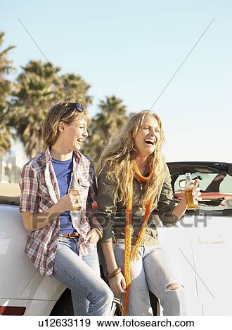 Stock Photo - Two women drinking beer next to convertible. Fotosearch