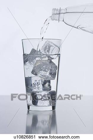 Stock Image - Water from bottle pouring into glass with ice cubes. Fotosearch