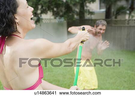Stock Image - Woman spraying man with hose in backyard. Fotosearch