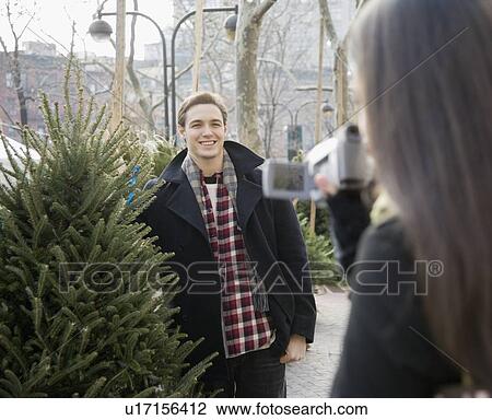 Stock Image - Woman video recording boyfriend with Christmas tree. Fotosearch