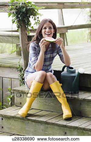 Stock Image - Woman wearing rubber boots and eating watermelon on deck. Fotosearch