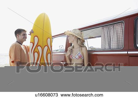Stock Image - Young couple standing with surfboard by van on beach. Fotosearch