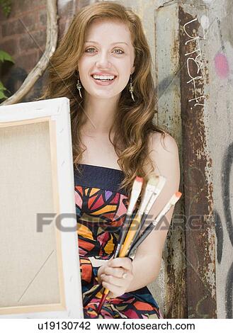 Stock Image - Young woman holding paintbrushes and canvas. Fotosearch