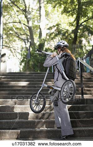 Stock Image - Businesswoman Carrying Bike on Steps. Fotosearch