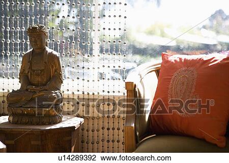 Stock Image - Buddha statue on end table near armchair. Fotosearch