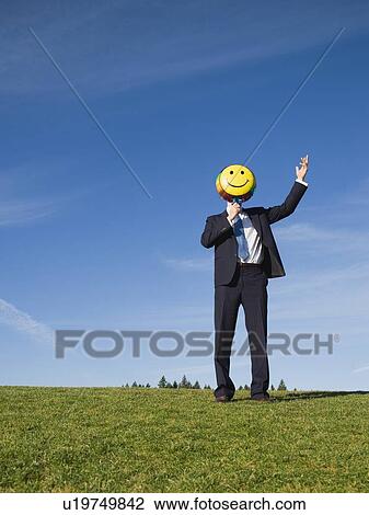 Stock Image - Businessman holding smiley face balloon over face. Fotosearch