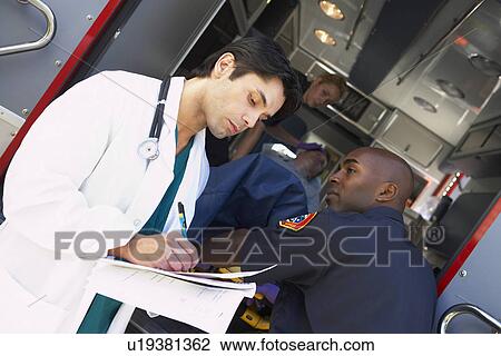 Stock Image - Hospital doctor taking notes as paramedics arrive with patient. Fotosearch