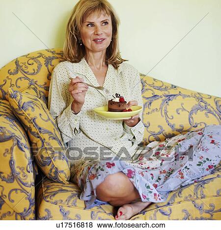 Stock Photo - Mature woman sitting on a couch eating cake. Fotosearch