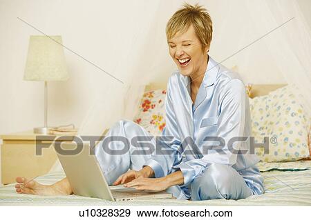 Stock Photo - Mature woman using a laptop on the bed and laughing. Fotosearch