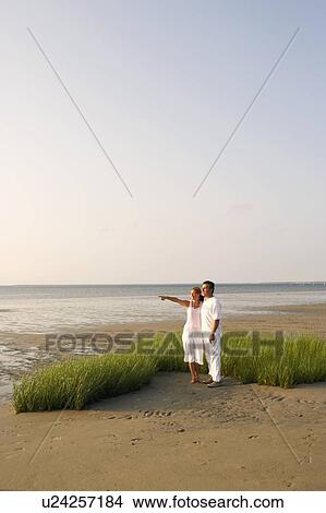 Picture - Mid adult woman pointing forward with a mature man standing beside her on the beach. Fotosearch