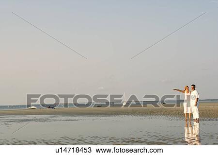 Stock Image - Mid adult woman pointing forward with a mature man standing beside her on the beach. Fotosearch