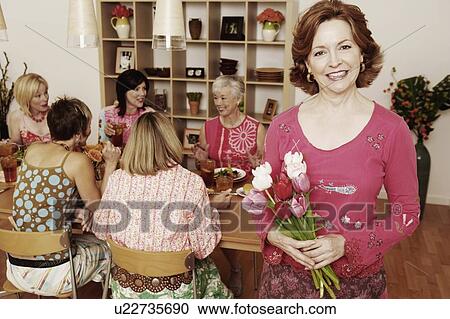 Portrait of a mature woman holding a bouquet of flowers and her friends sitting at the dining table in the background View Large Photo Image Stock Image - Portrait of a mature woman holding a bouquet of flowers and her friends sitting at the dining table in the background. Fotosearch