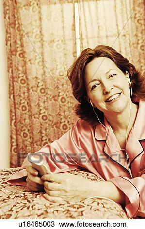 Stock Image - Portrait of a mature woman leaning against the bed and listening to an MP3 player. Fotosearch