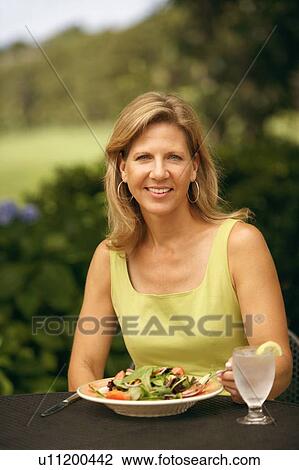 Stock Image - Portrait of a mature woman sitting at the table with a plate of vegetable salad in front of her. Fotosearch