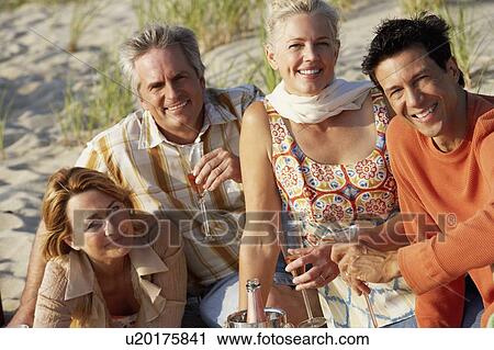 Stock Image - Portrait of two mature couples sitting on the beach and holding champagne flutes. Fotosearch