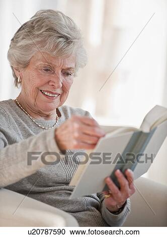 Stock Image - Senior woman reading book on sofa. Fotosearch
