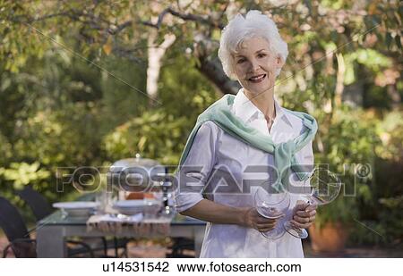 Stock Image - Senior woman standing in garden holding wineglasses. Fotosearch