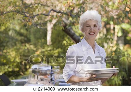 Stock Photo - Senior woman standing in garden holding stack of plates. Fotosearch