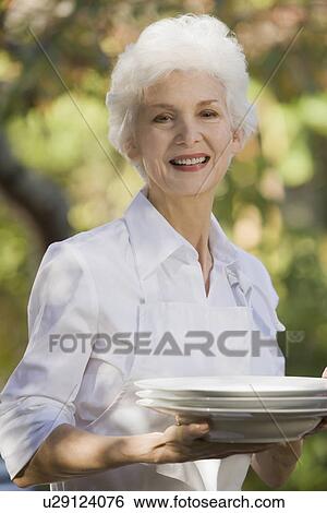 Stock Photograph - Senior woman standing in garden holding stack of plates. Fotosearch