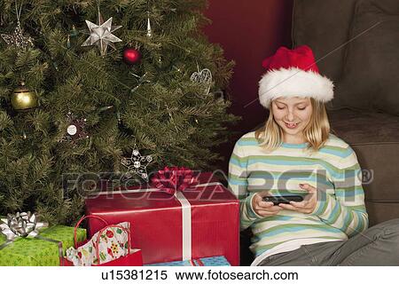 Stock Photography - Teenage girl (13-15) sitting in front of Christmas tree and text messaging. Fotosearch