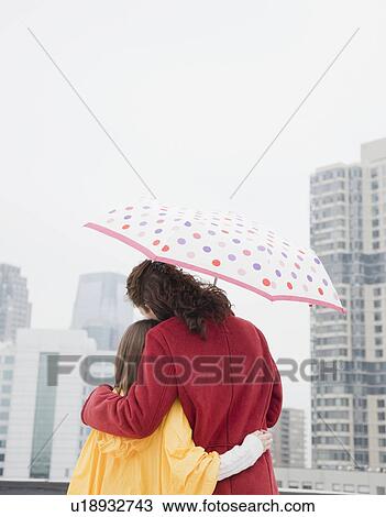 Stock Image - "USA, New York City, mother and daughter (10-12 years) embracing under umbrella, rear view, Jersey City, New Jersey". Fotosearch