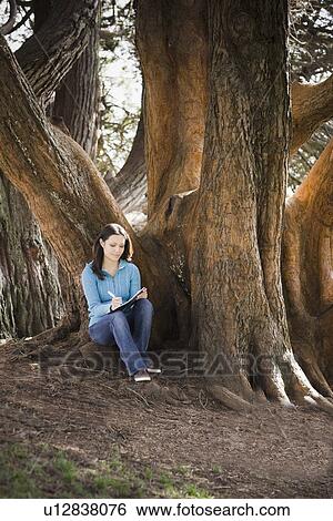 Stock Photograph - Woman writing diary by tree in park. Fotosearch