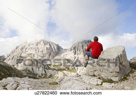 Stock Image - Man sitting on rocks looking at peaks. Fotosearch