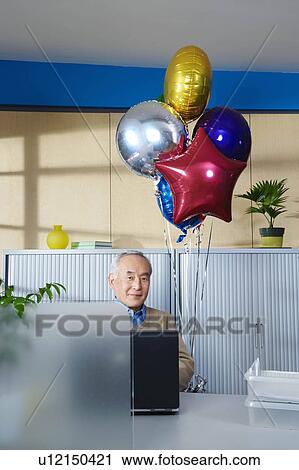 Stock Image - Senior man with balloons at office desk. Fotosearch