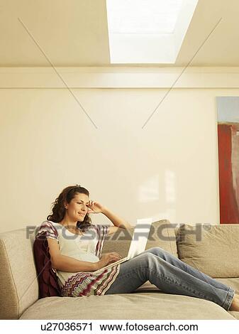 Stock Image - Woman sitting on sofa using laptop. Fotosearch