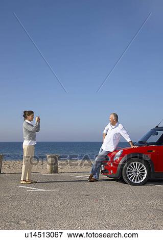 Stock Photo - Woman taking photo of man by car. Fotosearch