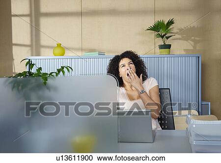 Stock Image - Woman yawning at desk in office. Fotosearch