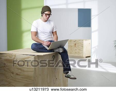 Stock Image - Young man sitting on box with laptop. Fotosearch