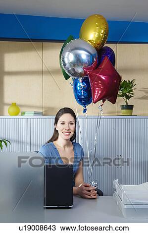 Stock Image - Young woman with balloons at desk. Fotosearch