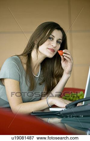 Stock Photo - Businesswoman eating a strawberry at laptop. Fotosearch