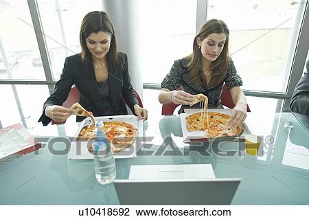 Stock Image - Businesswomen eating pizza at desk. Fotosearch