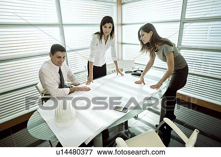 Professionals looking at architectural plans on desk View Large Photo Image Stock Photo - Professionals looking at architectural plans on desk. Fotosearch