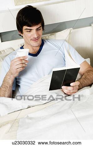 Stock Image - Young man in bed with book and coffee cup. Fotosearch