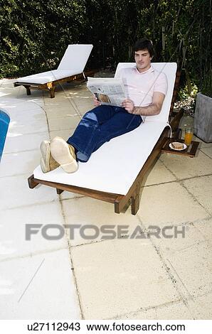 Stock Image - Young man on chaise lounge by pool reading. Fotosearch