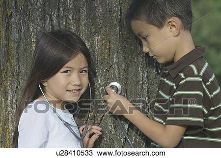 Stock Image - Five year old sister and seven year old brother with stethescope on tree, Winnipeg, Canada. Fotosearch