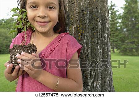 Stock Photography - Seven year old girl holding seedling beside tree, Winnipeg, Canada. Fotosearch