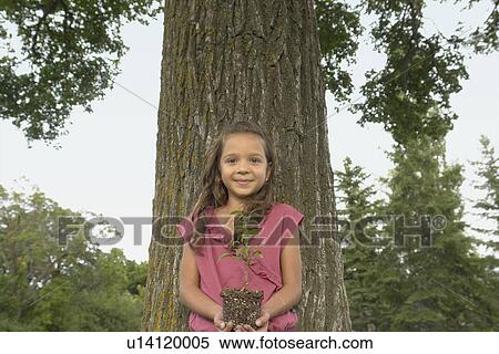 Stock Photography - Seven year old girl holding seedling beside tree, Winnipeg, Canada. Fotosearch