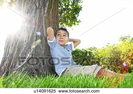 Stock Photography - Six year old boy seated beside large tree, Winnipeg, Canada. Fotosearch