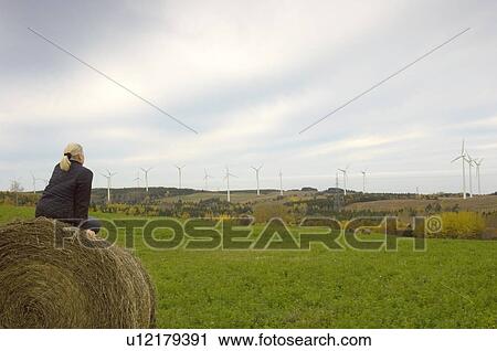 Stock Image - Woman on hay bale looking at wind turbines, St. Lawrence River, Cap-Chat, Quebec. Fotosearch