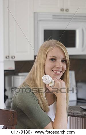 Stock Photo - Young woman holding compact fluorescent bulb in kitchen, Brandon, Canada. Fotosearch