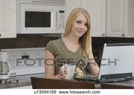 Stock Photo - Young woman in front of laptop computer in kitchen, Brandon, Canada. Fotosearch