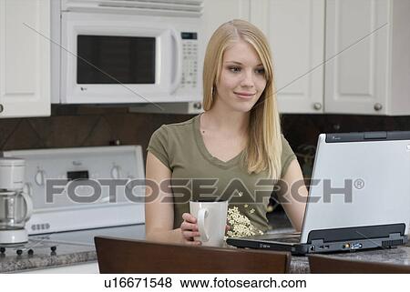 Stock Photo - Young woman in front of laptop computer in kitchen, Brandon, Canada. Fotosearch